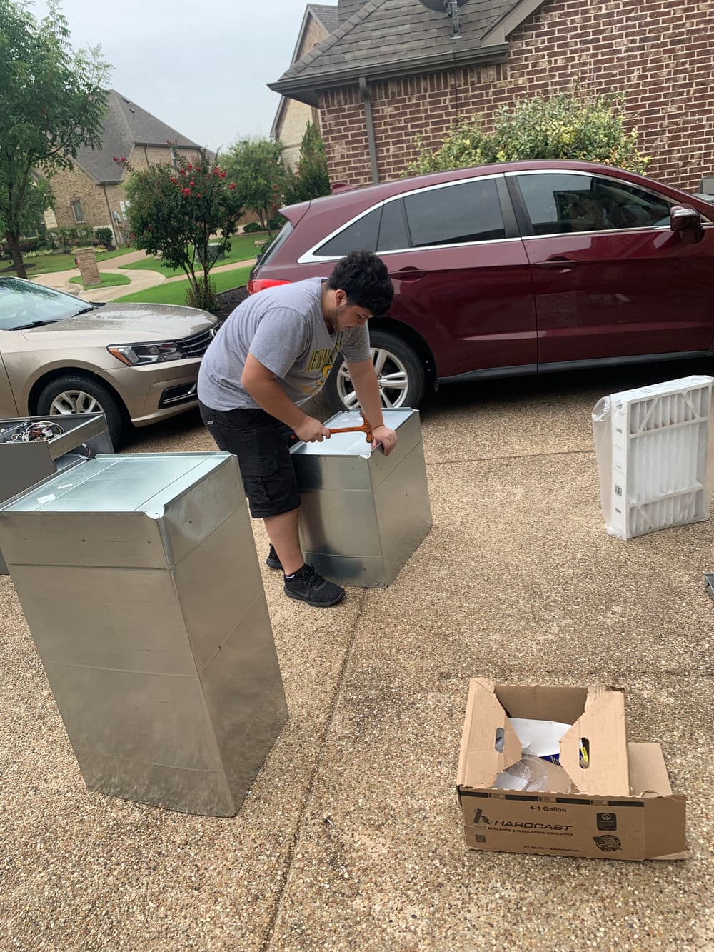 Teen assembling metal boxes in driveway with car and cardboard box in background.